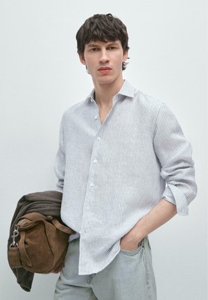 Junger Mann, der ein weißes und grau gestreiftes Hemd sowie helle Jeans trägt, hält vor einem schlichten Hintergrund eine braune Wildledertasche und eine Jacke.