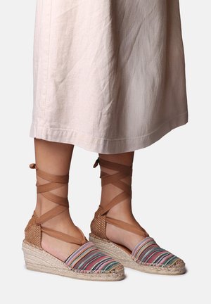 Keil-Espadrilles mit mehrfarbig gestreiftem Stoff und braunen Bändern, die um den Knöchel gewickelt sind, getragen zu einem hellbeigen Midirock.