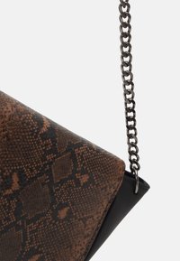 Hnědá a černá crossbody taška s hadím vzorem na bílém pozadí, s tlustým stříbrným řetízkovým popruhem.