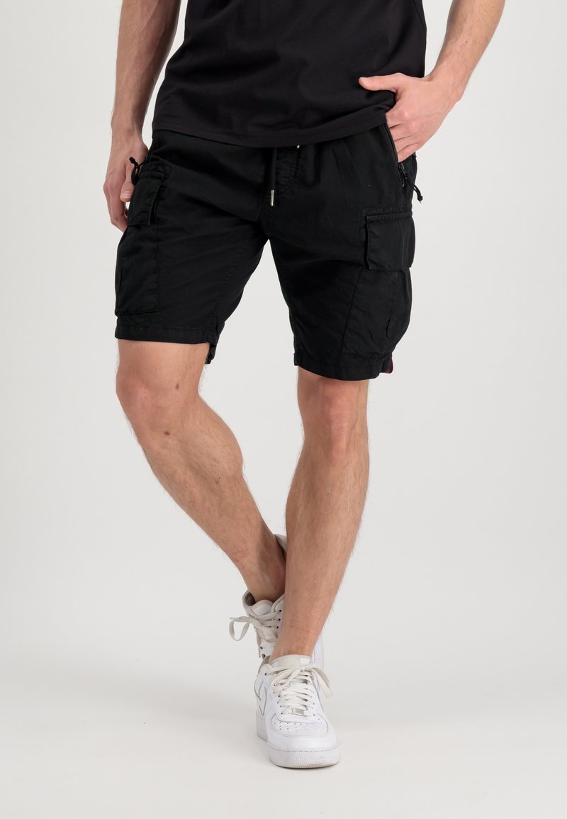 Pantaloni cargo neri con multiple tasche, vita elastica e chiusura con coulisse. Il tessuto ha una texture liscia. Indossati con sneakers bianche.