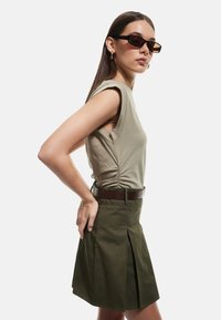 Gonna plissettata verde oliva con una cintura marrone, abbinata a un top senza maniche color khaki chiaro con arricciature laterali. Occhiali da sole scuri completano l'outfit.