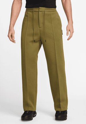 Homme portant un pantalon large vert olive avec coutures avant et taille à cordon, associé à des chaussures noires et un haut noir.