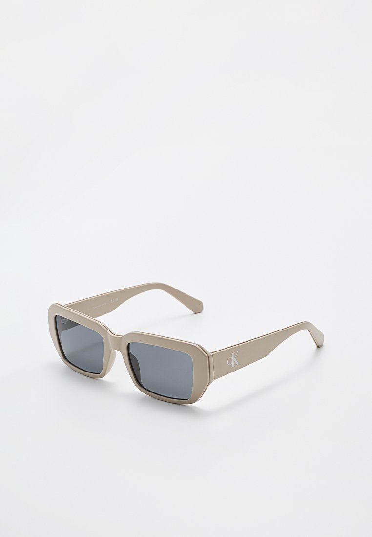 Beige rechteckige Sonnenbrille mit dunklen Gläsern und "cK"-Logo an den Bügeln, die auf einer weißen Oberfläche liegen.