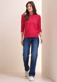 Röd stickad huvtröja med en ficka fram och dragsko, i kombination med blå avslappnade jeans och vita sneakers. Slät textur och avslappnad design.