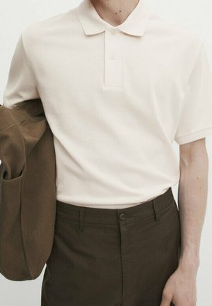 Personne portant un polo blanc à manches courtes rentré dans un pantalon marron foncé, tenant une veste marron assortie sur un bras.