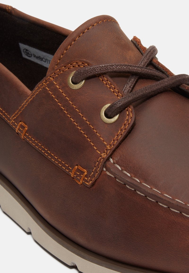 Chaussure de bateau en cuir marron avec des accents cousus, des œillets dorés et une semelle beige. Surface texturée et lacets marron foncé.