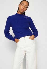 Royal blue ribbed sweater with a high neck and button accents on the shoulder, paired with white wide-leg pants.