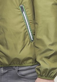 Olivgrön bomberjacka med en slät yta, försedd med en sidoficka med vita dragkedjor och gröna detaljer längs kanterna.
