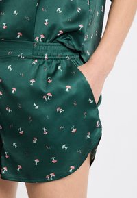 Grüne Satin-Shorts mit einem Muster aus roten Pilzen und kleinen Blumen. Der Stoff hat eine glatte Textur und einen elastischen Bund mit Taschen.