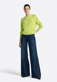 Limoengroen gebreide trui met ronde hals, gecombineerd met donkerblauwe denim jeans met wijde pijpen. De trui heeft geribbelde manchetten en zoom.