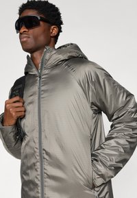 Szürke kapucnis dzseki, fényes, könnyű anyagból készült; cipzáras záródás, oldalsó zsebek és elegáns szabás jellemzi. Fekete napszemüveg kontrasztot ad.