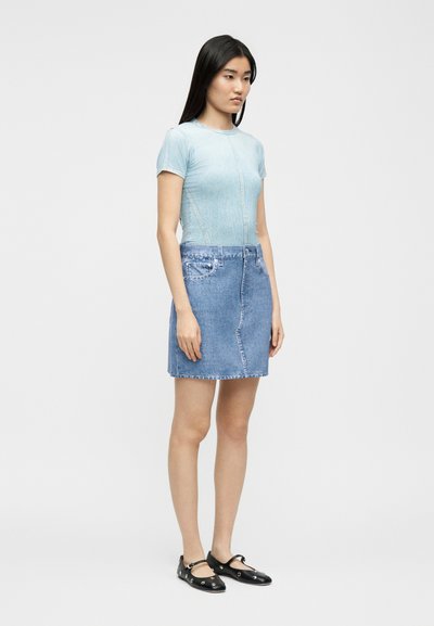 Νεαρή γυναίκα στέκεται, φορώντας ένα ανοιχτό μπλε εφαρμοστό μπλουζάκι, τζιν φούστα με τσέπες και μαύρα επίπεδα παπούτσια με λουράκια και ασημένιες τρύπες.