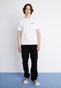 Camiseta de algodón blanca con un pequeño logotipo bordado en el pecho, combinada con pantalones cargo negros y zapatillas grises con detalles en verde.