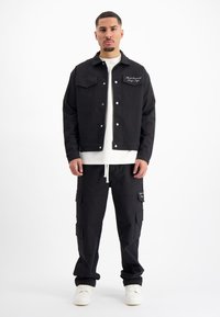 Zwarte spijkerjack met twee borstzakken, witte stiksels, gecombineerd met zwarte cargo broek met meerdere zakken en witte sneakers.