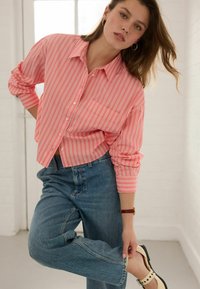 Roze en witte gestreepte button-up shirt met een zak, gedragen met hoog-waist blauwe jeans en witte schoenen met zwarte accenten.