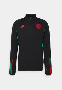 Camisa atlética de manga larga negra con media cremallera, logo rojo del Manchester United, paneles laterales verdes y detalles de rayas rojas y verdes en las mangas.