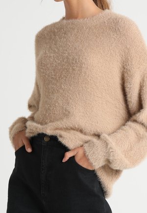 Beige flauschiger Pullover mit Rundhalsausschnitt, langen Ärmeln und gerippten Kanten, kombiniert mit hochgeschnittenen schwarzen Jeans. Weiche Textur sichtbar.