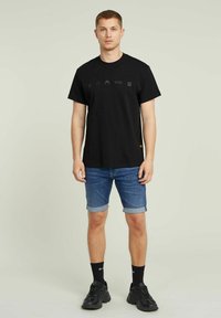 Tricou negru din bumbac cu pictograme grafice pe piept, asortat cu pantaloni scurți din denim rulați și adidași sport negri.