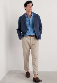 Blazer de lino azul marino, camisa a rayas azul claro, pantalones beige y mocasines trenzados marrones. Textura suave, corte casual y dobladillos de pantalón enrollados.