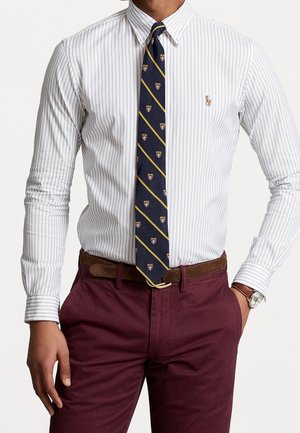 Homme portant une chemise blanche et gris clair à rayures avec boutons, une cravate bleu marine à motif, un pantalon bordeaux, une ceinture marron et une montre-bracelet.