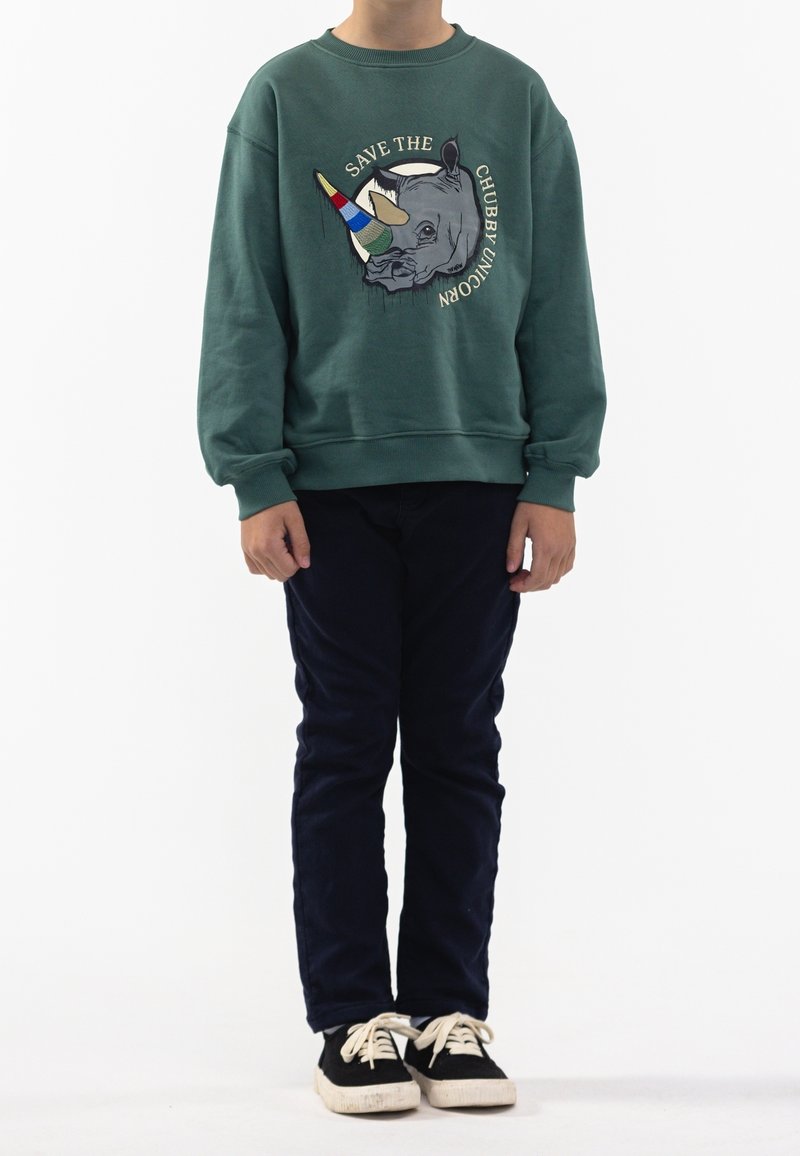 Sweatshirt vert avec un graphique de rhinocéros et le texte "SAUVEZ LA LICORNE RONDELETTE", associé à un pantalon bleu marine et des baskets noires.