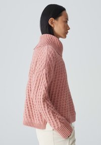 Rosa gestrickter Pullover mit hohem Kragen, der ein strukturiertes Muster und gerippte Bündchen aufweist, seitlich vor einem neutralen Hintergrund gezeigt.