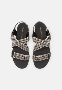 Mustat sandaalit, joissa on ristikkäiset kudotut hihnat, joissa on kerma- ja musta kuvio. Pohjat ovat pehmustetut ja sileäpintaiset. Brändäys näkyvissä.