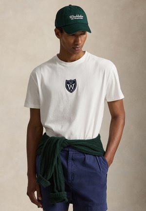 Homme portant un t-shirt blanc avec le logo de Wimbledon, un short bleu marine, un pull vert foncé noué autour de la taille et une casquette Wimbledon, regardant vers le bas.
