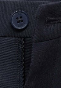 Gros plan sur un tissu bleu marine avec un bouton gravé du nom de la marque et une fermeture éclair en métal. Surface texturée avec des lignes diagonales.