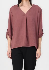Blusa leggera color rosa con scollo a V e maniche arrotolabili. Presenta una vestibilità rilassata e un tessuto testurizzato. Indossati sotto ci sono pantaloni neri.