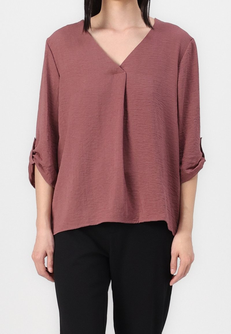 Blusa leggera color rosa con scollo a V e maniche arrotolabili. Presenta una vestibilità rilassata e un tessuto testurizzato. Indossati sotto ci sono pantaloni neri.