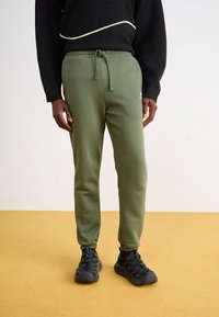 Grønne sweatshirtbukser med elastisk talje og snor. Mansjetter ved anklerne. Sammen med sorte sportssko. Simpelt design, glat tekstur.