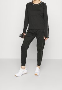 Svart långärmad träningströja med reflexsömmar, tillsammans med svarta tapered joggers. Vita träningsskor med gummisålar och en vattenflaska.