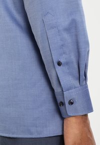 Blue patterned dress shirt with a textured weave, featuring a buttoned cuff and dark buttons, showing a close-up of the sleeve detail.