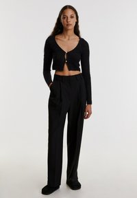 Schwarze, verkürzte Strickjacke mit einem Schlüssellochdetail und Knopfverschluss, kombiniert mit hoch taillierten, gefalteten schwarzen Hosen. Glatte Stoffstruktur.