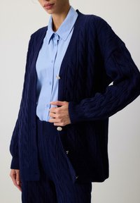 Marineblå kabelstrikket cardigan med lange ærmer, ribbede manchetter og dekorative knapper; parret med en lyseblå skjorte med krave under.