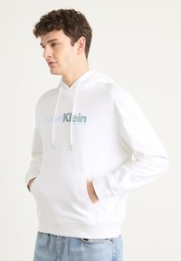Biała bawełniana bluza z kapturem ze sznurkiem, przednia kieszeń oraz logo Calvin Klein w jasnoniebieskim kolorze. Posiada prążkowane mankiety i dół. Luźny krój.
