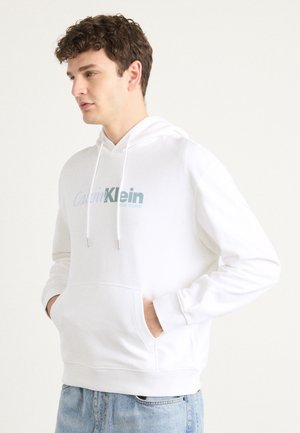 Hvit bomullhetter med snor, frontlomme og Calvin Klein-logo i lyseblå. Har ribbestrikkede mansjetter og kant. Løs passform.