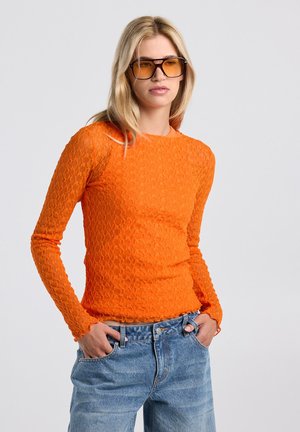 Jonge vrouw met een oranje gestructureerde top met lange mouwen, blauwe spijkerbroek en grote zonnebril met amberkleurige glazen, handen in de zakken, neutrale achtergrond.