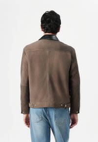 Brun ruskindsjakke med krave i sort læder, med synlige sømme og trykknapsdetaljer, båret over blå jeans.