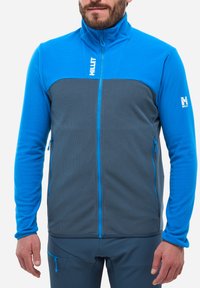 Veste en polaire bleue et grise avec un col haut, un corps texturé et des manches lisses. Présente des poches zippées et des détails de couture contrastants.