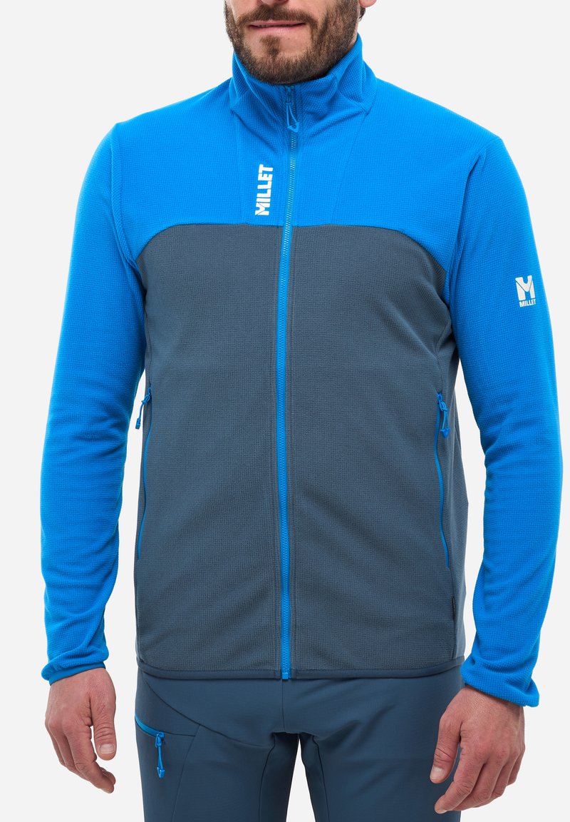 Veste en polaire bleue et grise avec un col haut, un corps texturé et des manches lisses. Présente des poches zippées et des détails de couture contrastants.