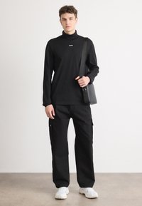 Svart langermet turtleneck med hvit logo, sammen med løse svarte cargo-bukser og hvite sneakers. En liten svart skulderveske bæres på tvers av kroppen.