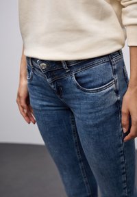 Jeans oscuros deslavados con una cintura de altura media, con bolsillos frontales, cierre de cremallera y un botón plateado, combinados con un suéter claro.