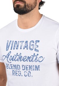 Bela majica s kratkimi rokavi iz bombaža s prikazom modre obrabljene tipografije: "VINTAGE Authentic Blend Denim REG. CO." na sprednji strani.