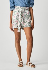 Bermudas florales en blanco con patrones en rosa y verde, con una cintura alta, cinturón anudado y dobladillos enrollados. Combinadas con sandalias ligeras.