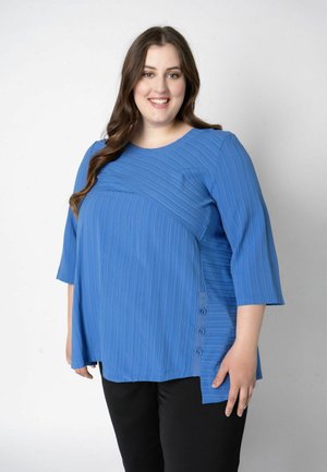 Femeie zâmbitoare cu păr lung și maro, purtând un top albastru texturat cu mâneci trei sferturi și pantaloni negri, stând în fața unui fundal deschis.