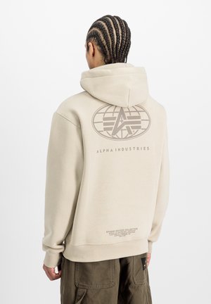 Persoon met vlechtjes die een beige Alpha Industries-hoodie draagt met logo en tekst op de achterkant, gecombineerd met olijfgroene cargobroek, met de rug naar de camera gekeerd.