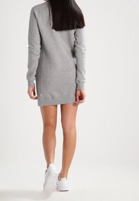 Graues Sweatshirtkleid mit langen Ärmeln, Rundhalsausschnitt und seitlichen Schlitzen. Weicher Stoff und knielanges Design. Kombiniert mit weißen Sportschuhen.