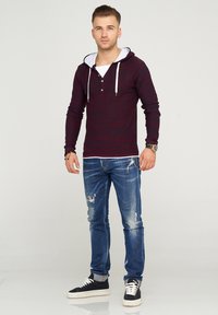 Bordeauxrot gestreifter Langarm-Hoodie mit weißem Innenfutter und Kordeln, kombiniert mit ausgewaschenen blauen Jeans und schwarzen Sneakers.